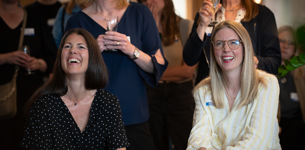 Tre kvinnor skrattar glatt med människor i bakgrunden.